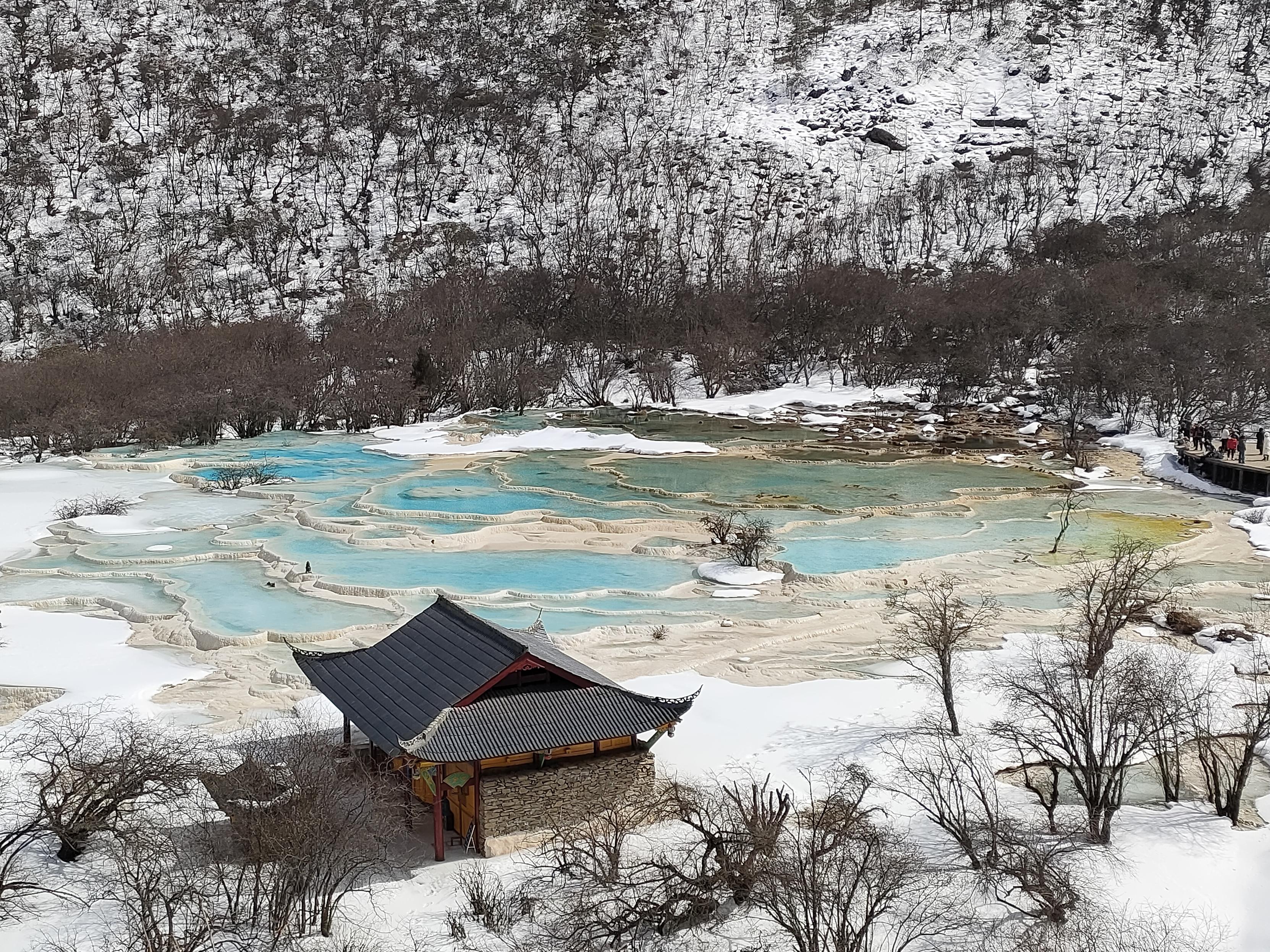 高角度拍摄的户外风景照片,展示了一个雪覆盖的山谷和一组石灰华池。照片中心是一组多层叠错的浅色石灰华池,池中蓄满了清澈的碧蓝色水。池水呈现出明亮的蓝色和绿色,池塘的形状不规则,由浅色的岩石边缘分隔开,形成一个个小型的水域。在照片的下方可看到一座传统的中国式建筑,屋顶呈深灰色,建筑的墙壁呈浅棕色。建筑周围覆盖着白雪和枯萎的树木。照片背景是覆盖着白雪的山坡,山坡上覆盖着稀疏的树木,树木呈深棕色和灰色,在雪地上投下细长的影子。山坡的轮廓清晰,呈现出锯齿状的线条。在照片的右侧可以看到一条小路,上面有一些游客在观赏风景。